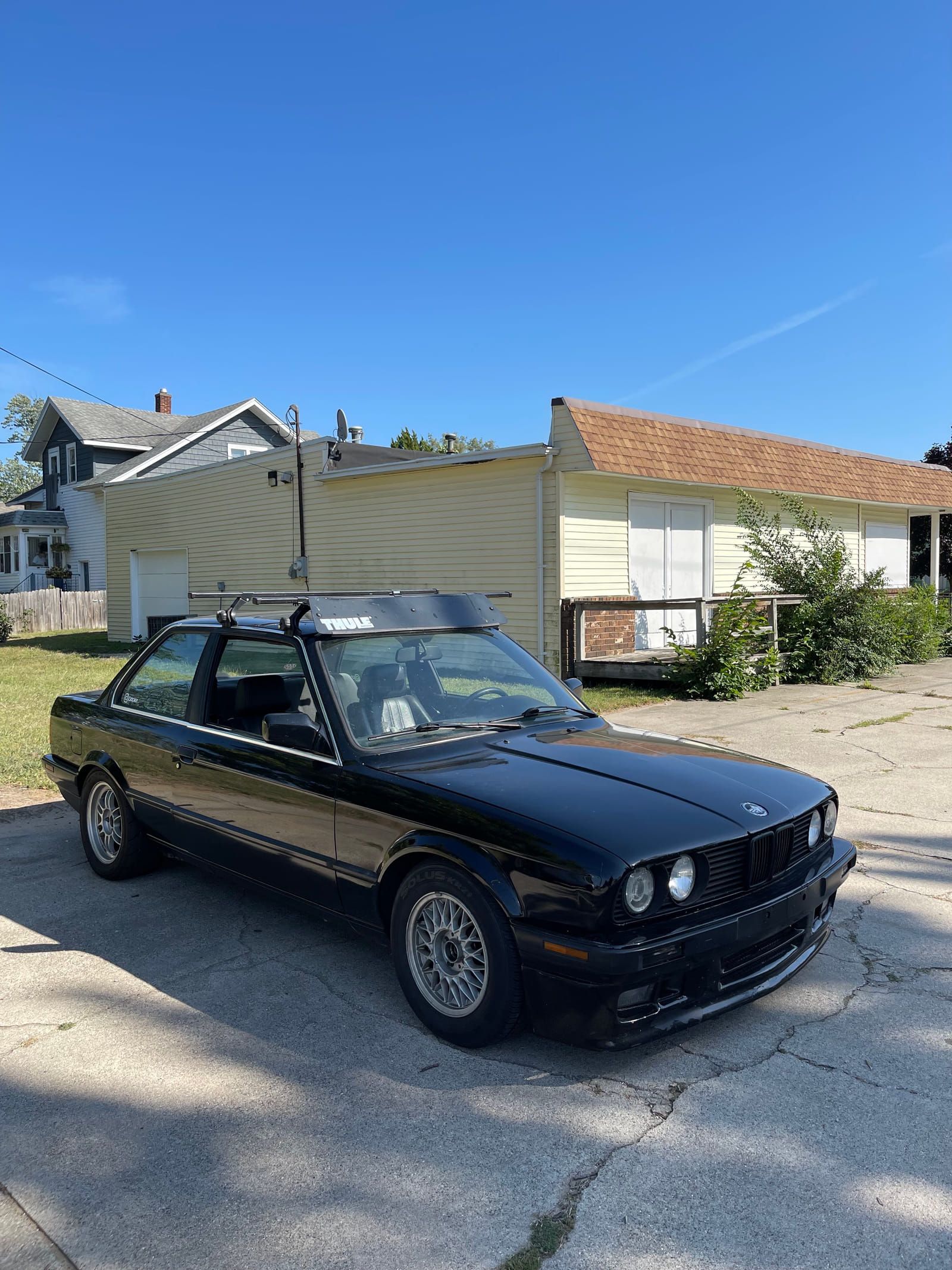 Black 1990 BMW 325i coupe with aftermarket wheels and a roof rack, parked on a driveway