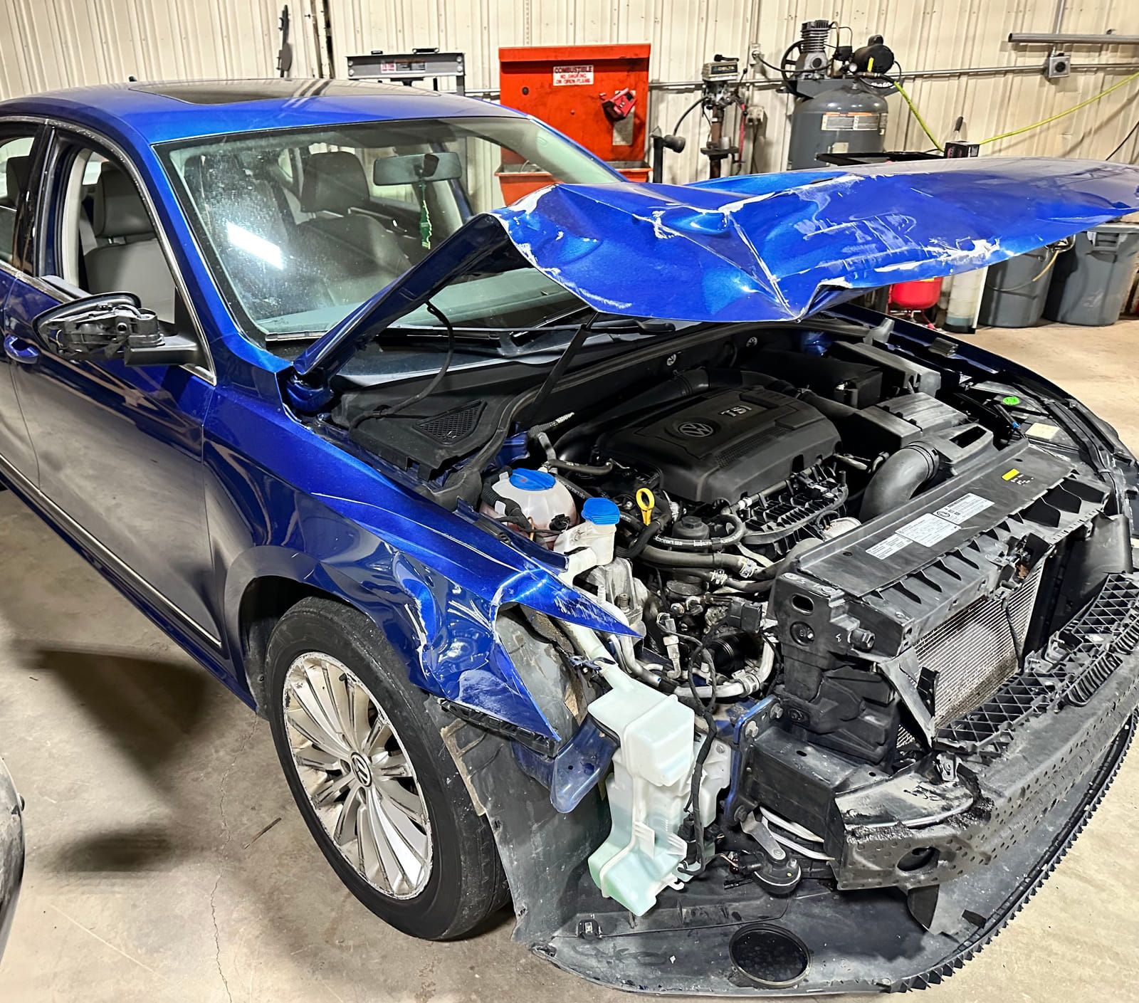 Blue 2014 Volkswagen Passat in a workshop with heavy front-end damage, hood folded and headlamp assemblies removed