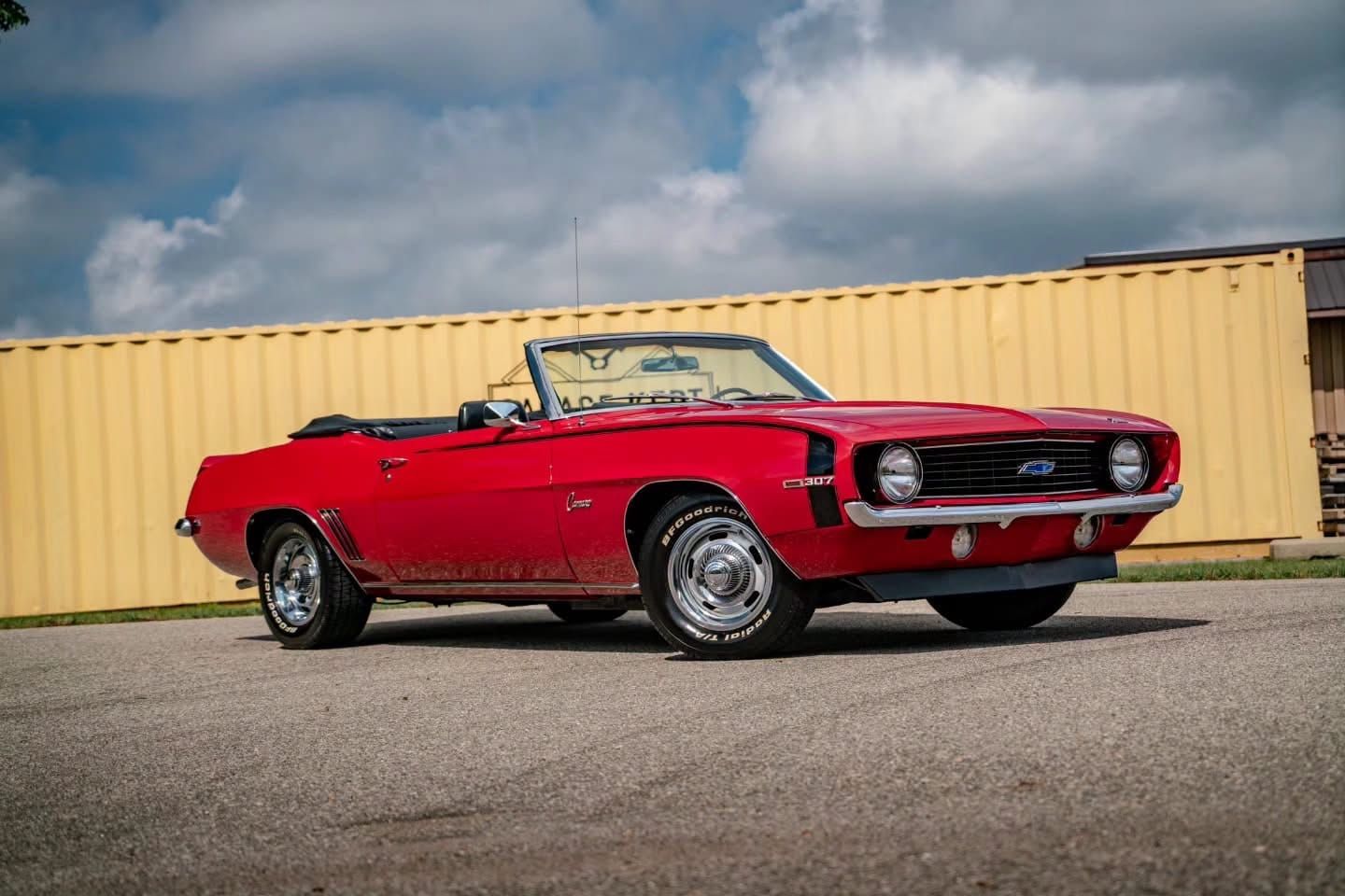Red 1969 Chevrolet Camaro convertible with the top down, parked outside a tan metal building